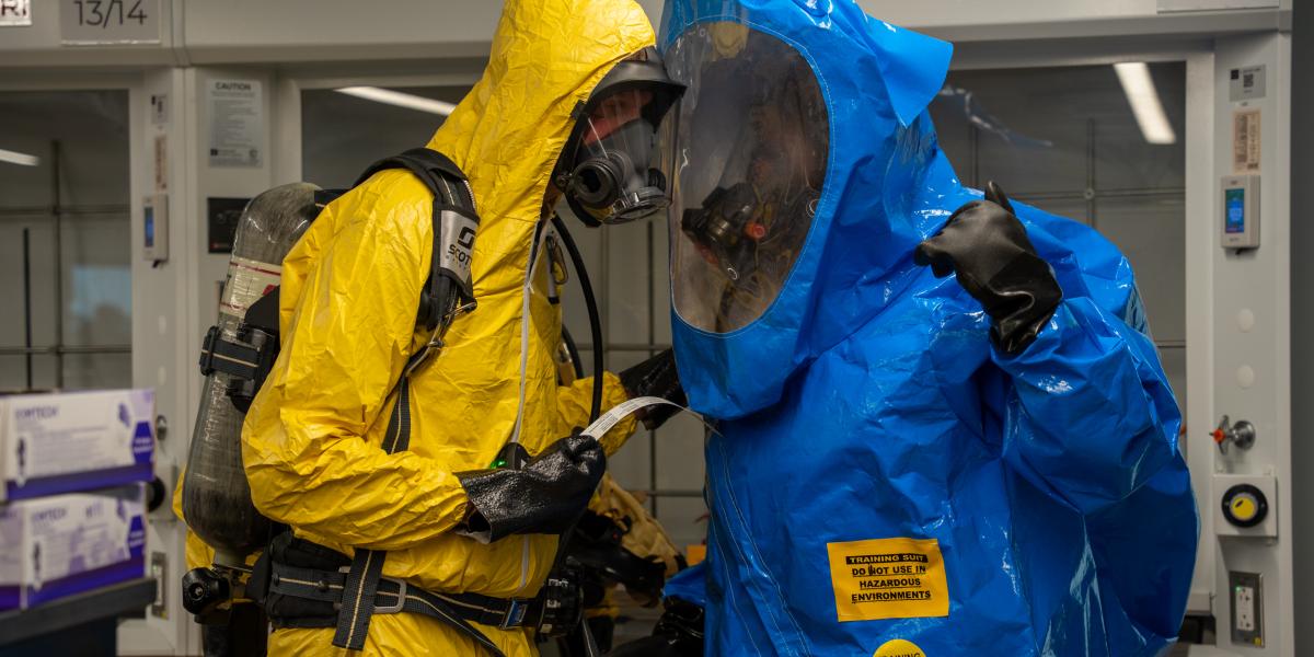 Two people in yellow and blue HAZMAT suits are seen speaking to each other.