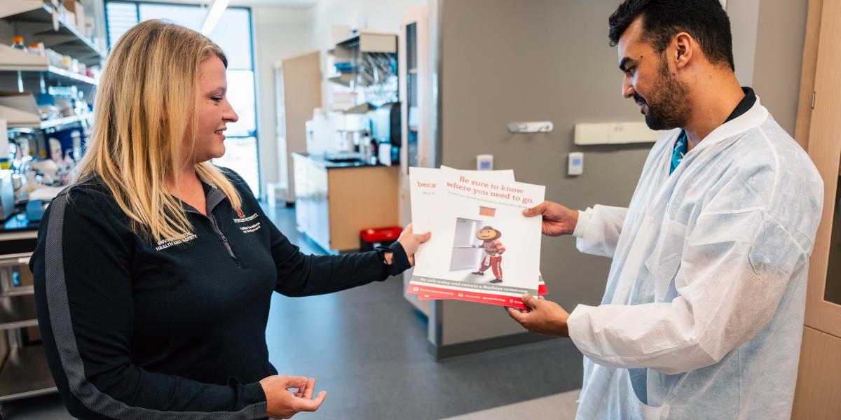 Jenna Dickerson is seen giving a student researcher a poster with Brutus Buckeye on the front that says, "Be safe today remain a Buckeye tomorrow"