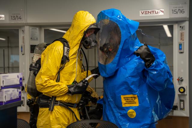 Two people in yellow and blue HAZMAT suits are seen speaking to each other.
