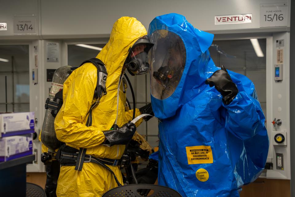 Two people in yellow and blue HAZMAT suits are seen speaking to each other.