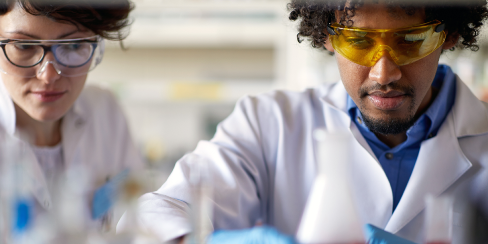 two people with lab googles, lab coats and gloves